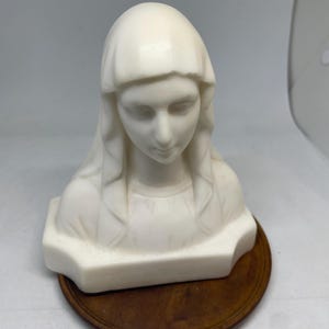 Op de afbeelding: Een crèmekleurige buste-sculptuur van een vrouw met een gedrapeerde hoofdbedekking. De sculptuur is gemonteerd op een ronde, donkerbruine houten basis. Het gezicht van de vrouw is sereen, met neergeslagen ogen.
