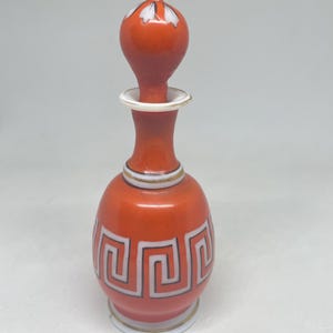 Puede incluir: Un pequeño frasco de perfume de cerámica naranja con tapón a juego. El frasco presenta un patrón de greca blanca alrededor del cuerpo, acentuado con finas líneas negras. El tapón tiene una parte superior redondeada con detalles blancos.