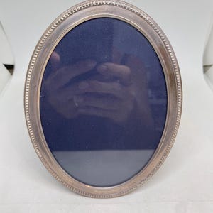 May include: An oval-shaped picture frame with a metallic silver finish and a beaded texture along the outer edge. The frame features a dark navy blue backing material inside the oval opening, creating a contrast with the metallic frame border.