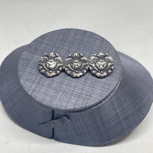 Op de afbeelding: Een set van drie cirkelvormige zilverkleurige metalen knopen of decoratieve accenten, elk voorzien van een gedetailleerd reliëf bloem- of bloembladontwerp met een donkere, geoxideerde afwerking in de uitsparingen om de patronen te benadrukken.