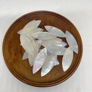 May include: A collection of iridescent, oval-shaped mother-of-pearl pieces with etched designs, displayed in a round, wooden bowl. The pieces have a pearlescent sheen, reflecting various colors. The bowl is a warm, medium brown.