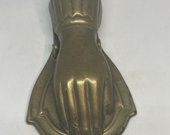 May include: A vintage brass hand-shaped clip, likely for holding papers or notes. The clip features a detailed hand design with a decorative base. The brass has a warm, aged patina, suggesting it is an antique or vintage item.
