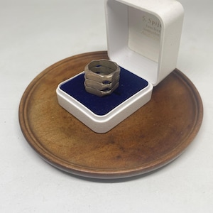 Op de afbeelding: Een zilverkleurige ring met een gestapeld ontwerp van meerdere banden met uitgesneden details. De ring wordt gepresenteerd in een wit juwelendoosje met een donkerblauwe fluwelen voering, rustend op een ronde houten basis.