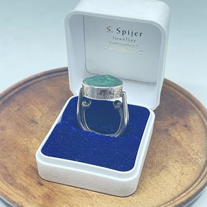 May include: A silver ring with a textured green stone inlay, presented in a white hinged jewellery box with a blue velvet interior. The box lid features the printed text S. Spijer Juwelier Koningsplein 1 Amsterdam-C in black and gold lettering.