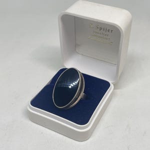 Könnte beinhalten: Ein Silberring mit einem großen, glatten, ovalen schwarzen Stein, präsentiert in einer weißen aufklappbaren Schmuckschatulle mit blauem Samtinnenfutter. Der Innendeckel der Schatulle zeigt den Text S. Spijer, Juwelier, Amsterdamsestraatweg 1, Utrecht-C.