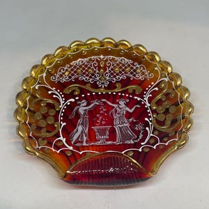 Può includere: Un piatto decorativo a forma di conchiglia con una sfumatura di colore dal rosso all'arancione. Il bordo è dorato e il centro presenta un'illustrazione bianca di due figure. Il piatto ha un bordo smerlato ed è probabilmente in vetro.