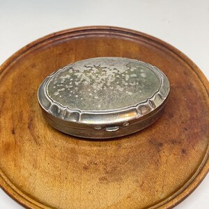 Puede incluir: Una caja ovalada de plata antigua con un borde decorativo, sobre una bandeja redonda de madera. La caja tiene una superficie deslustrada y texturizada. La bandeja de madera tiene un tono marrón cálido.