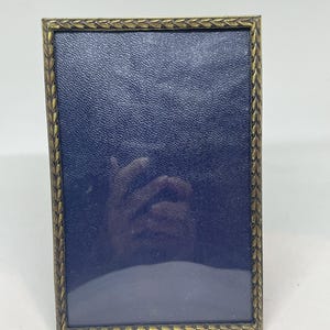 May include: Rectangular antique brass picture frame with a textured navy blue backing. The frame has a decorative leaf pattern around the edges. The frame is designed to hold a photograph or artwork.