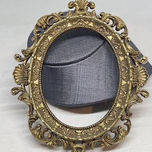 May include: An oval-shaped picture frame with a metallic gold finish. The frame features intricate scrollwork, shell-like motifs on the sides, and a fleur-de-lis design at the top and bottom. Small white beads are embedded along the inner border.