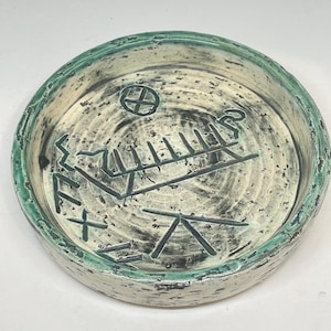 Puede incluir: Un plato redondo de cerámica con una superficie texturizada y un borde verde turquesa. El interior presenta marcas y símbolos abstractos verde oscuro sobre un fondo crema. El plato tiene un aspecto rústico y hecho a mano.