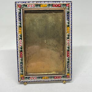 May include: A rectangular picture frame with a mosaic border of red, yellow, and white flowers on a blue background. The frame has a gold-coloured interior and rests on small, gold-coloured feet.