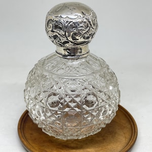 May include: A clear glass perfume bottle with a spherical body featuring a diamond-cut pattern. The bottle is topped with a round, ornate silver-colored metal stopper that displays intricate embossed floral and scrollwork designs throughout.