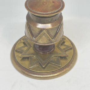 Pode incluir: Tinheiro antigo de latão com um desenho de estrela na base e padrões geométricos decorativos. O tinteiro tem um topo cor de cobre e um corpo cilíndrico. A base é circular e tem uma borda elevada.