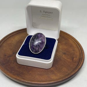 Op de afbeelding: Een zilveren ring met een grote, ovaalvormige paars en zwart gemarmerde steen, gepresenteerd in een wit sieradendoosje met een blauwe fluwelen binnenkant. Het deksel bevat de tekst S. Spijer Juwelier Koningsplein 1 Amsterdam-C.