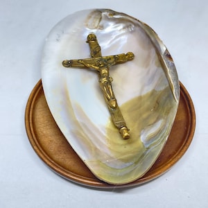 Pode incluir: Um crucifixo de metal dourado com a figura de Jesus, repousando dentro de uma grande concha de madrepérola iridescente. A concha está posicionada sobre uma base circular de madeira castanho-clara, lisa e com um padrão de veios visível.