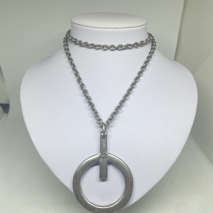 May include: A silver-toned necklace with a large circular pendant and a smaller chain that sits higher on the neck. The pendant has a vertical bar attached to the circle. The necklace is displayed on a white mannequin.