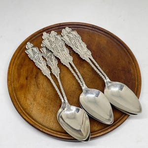 May include: Four silver spoons with ornate handles and oval bowls rest on a round, wooden plate. The spoons have a twisted design on the handles and decorative detailing at the top. The wooden plate is a warm brown color.