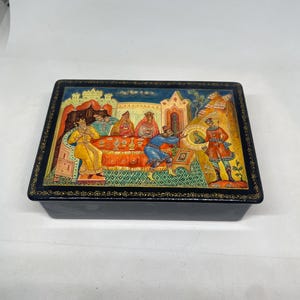 Puede incluir: Una caja lacada en negro de forma rectangular. La tapa presenta una pintura detallada y colorida que representa una escena histórica con figuras vestidas con trajes tradicionales. La caja está adornada con detalles dorados.