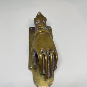 Puede incluir: Un llamador de puerta de latón con forma de mano con un puño en la muñeca, montado sobre una placa rectangular con extremos redondeados. El metal tiene un acabado pulido con una pátina oscura visible en los huecos de los dedos y el puño.