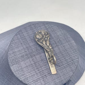 Könnte beinhalten: Eine silberfarbene Krawattenklammer aus Metall mit einem verzierten, geprägten Blumenmuster im oberen Bereich. Das Design umfasst einen abgerundeten, dekorativen Kopf mit komplexen Schnörkeln und eine glatte, gerade untere Leiste mit einer kleinen Öse.