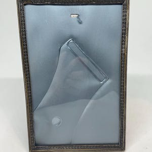 May include: A rectangular, antique-style picture frame with a decorative border. The frame is a dark metallic colour, possibly bronze or brass. The backing is a light blue fabric, and the stand is clear plastic.