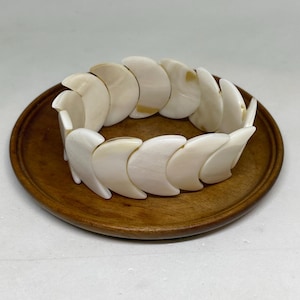 May include: A cream-colored bracelet with curved, shell-like segments. The bracelet is resting on a round, wooden plate. The segments are connected, forming a flexible band. The bracelet's design is organic and natural.