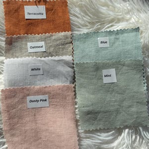 May include: A collection of fabric swatches in various colors, including terracotta, oatmeal, white, dusty pink, blue, and mint. Each swatch has a label with its color name. The swatches are arranged on a fluffy, white surface.