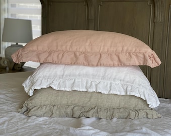 Puede incluir: Tres fundas de almohada con volantes en tonos rosa, blanco y beige apiladas sobre una cama blanca. Las almohadas están sobre una cama con cabecero de madera. La habitación tiene una paleta de colores neutros.