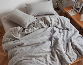 May include: A bed with light gray linen bedding, including a duvet cover and two pillowcases. The bedding is neatly arranged, with soft folds and a relaxed appearance. The bed is made of wood.