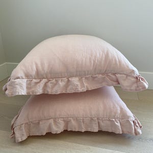 May include: Two light pink linen pillowcases stacked on top of each other. Each pillowcase has a ruffled edge. The pillows are a soft, muted pink color and appear to be made of a lightweight fabric.