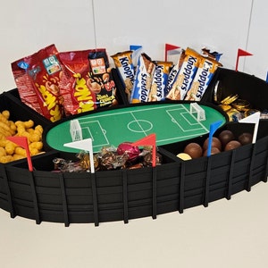 Può includere: Una ciotola per snack a forma di stadio in bianco e nero con bandiere rosso, bianco e blu. La ciotola è piena di vari snack, tra cui patatine, cioccolato e barrette di cioccolato. Le barrette di cioccolato sono etichettate "Knoppers".