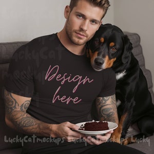 Puede incluir: Un hombre con una camiseta negra con el texto "Design here" en letras rosas está sentado en un sofá con un perro negro. Está sosteniendo un plato con un trozo de pastel de chocolate.