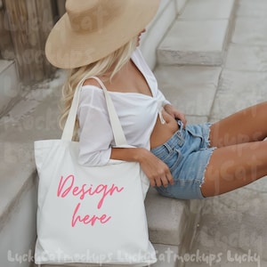 May include: A white canvas tote bag with the words "Design here" in pink lettering. The bag is being held by a person wearing a white shirt, denim shorts, and a straw hat.