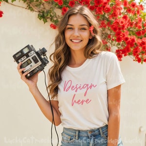 May include: A white t-shirt with the text "Design here" in pink lettering. The shirt is being held by a person with long brown hair and a flower in their hair. The person is standing in front of a white wall with red flowers.