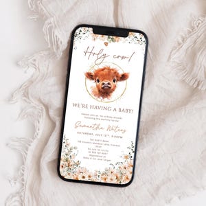 May include: A smartphone displays a baby shower invitation with a watercolor illustration of a Highland cow. The text reads "Holy cow! We're having a baby!" with event details and floral accents. The phone is set against a white, textured background.