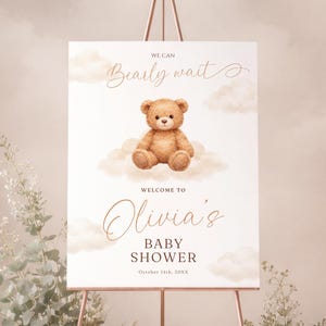 Peut inclure: Panneau de bienvenue pour une baby shower avec une illustration d'ours en peluche. Le panneau indique "We can bearly wait" et "Welcome to Olivia's Baby Shower". Le panneau est sur un chevalet en or rose, avec de la verdure et un ours en peluche.