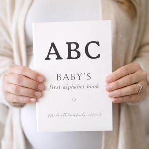 Puede incluir: Un libro de alfabeto para bebés blanco con la palabra "ABC" en letras negras grandes y en negrita. El texto "BABY'S first alphabet book" está debajo, con un pequeño símbolo de corazón. La portada del libro también incluye la frase "Made with love by family and friends."