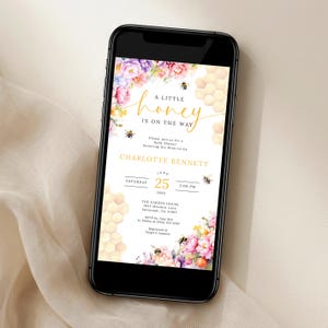 Peut inclure: Un smartphone affiche une invitation à une baby shower. Le design présente des arrangements floraux roses, violets et jaunes, des motifs en nid d'abeille et des illustrations d'abeilles. Le texte dit "A Little Honey is on the Way" avec les détails de l'événement.