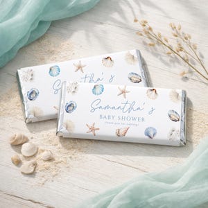 Peut inclure: Deux barres de chocolat avec des emballages blancs, décorées de coquillages, d'étoiles de mer et de coraux à l'aquarelle. Le texte "Baby Shower de Samantha" est affiché en écriture bleue. Les barres sont sur du sable avec des coquillages et un tissu bleu.