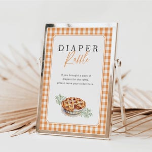 May include: A silver framed sign with a gingham orange border. The sign reads "DIAPER Raffle" in black and orange script. Below the title is text that reads "If you brought a pack of diapers for the raffle, please leave your ticket here." A watercolor pie illustration is at the bottom.