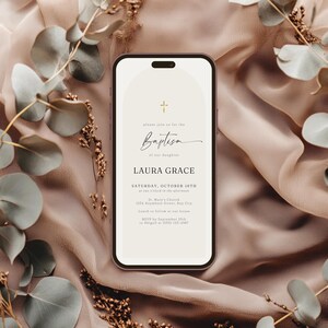 May include: A smartphone displays a baptism invitation with a cross icon and the word "Baptism" in elegant script. The invitation includes the name "LAURA GRACE" and event details. The phone rests on a textured, neutral-toned fabric, adorned with eucalyptus branches.