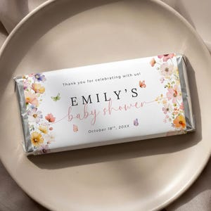 Puede incluir: Envoltorio rectangular de barra de chocolate con diseño floral y el texto "Baby shower de Emily". El envoltorio presenta flores rosas, naranjas y amarillas, con mariposas. También se incluye el texto "¡Gracias por celebrar con nosotros!"