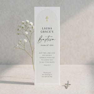May include: A white baptism card with gold cross and text. The card reads "LAURA GRACE'S Baptism October 16th 20XX" and a bible verse. A small silver cross is at the bottom.