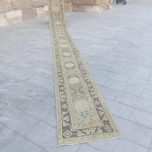 Può includere: Un lungo e stretto tappeto a passatoia caratterizzato da una palette di colori beige sbiadito e marrone scuro con intricati motivi geometrici e floreali. Il tappeto presenta un medaglione centrale, un bordo decorativo e frange corte.