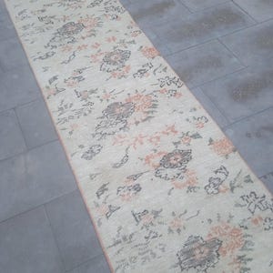 May include: A long, rectangular rug with a cream background and a floral pattern in shades of pink and black. The rug has a distressed look, giving it a vintage feel.