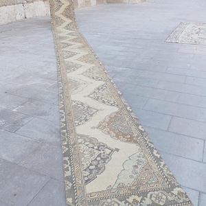 Puede incluir: Una alfombra de pasillo larga y estrecha con un patrón geométrico y floral repetitivo en tonos apagados de beige, gris, marrón y azul suave. La alfombra presenta un diseño central en zigzag flanqueado por medallones ornamentados y un borde detallado.