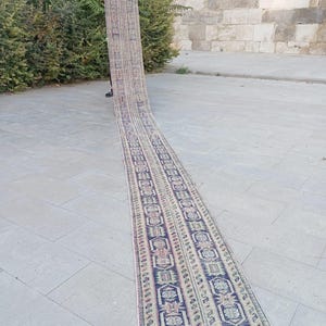 Peut inclure: Un long tapis de couloir rectangulaire avec un motif géométrique détaillé dans des tons de bleu, beige et rouge. Le tapis est présenté à l'extérieur, mettant en valeur sa longueur et son design complexe. Les bords du tapis ont des franges.