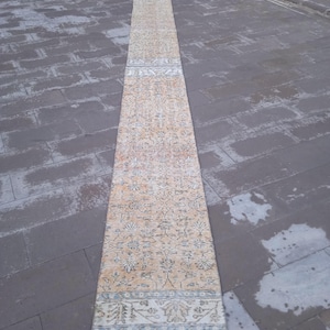 May include: A long, narrow, beige rug with a floral pattern and a blue and white border. The rug is laid out on a grey tiled floor.