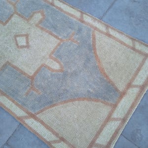 May include: A vintage rug with a geometric pattern in shades of beige, brown, and blue. The rug has a fringed edge and is made of a woven material.