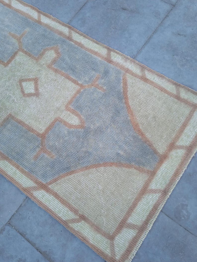 May include: A vintage rug with a geometric pattern in shades of beige, brown, and blue. The rug has a fringed edge and is made of a woven material.